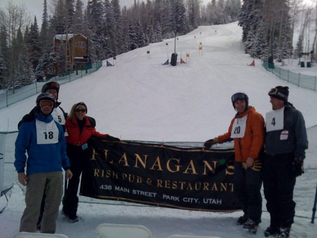 five people posing with a Flanagan's banner at the top of a ski slope